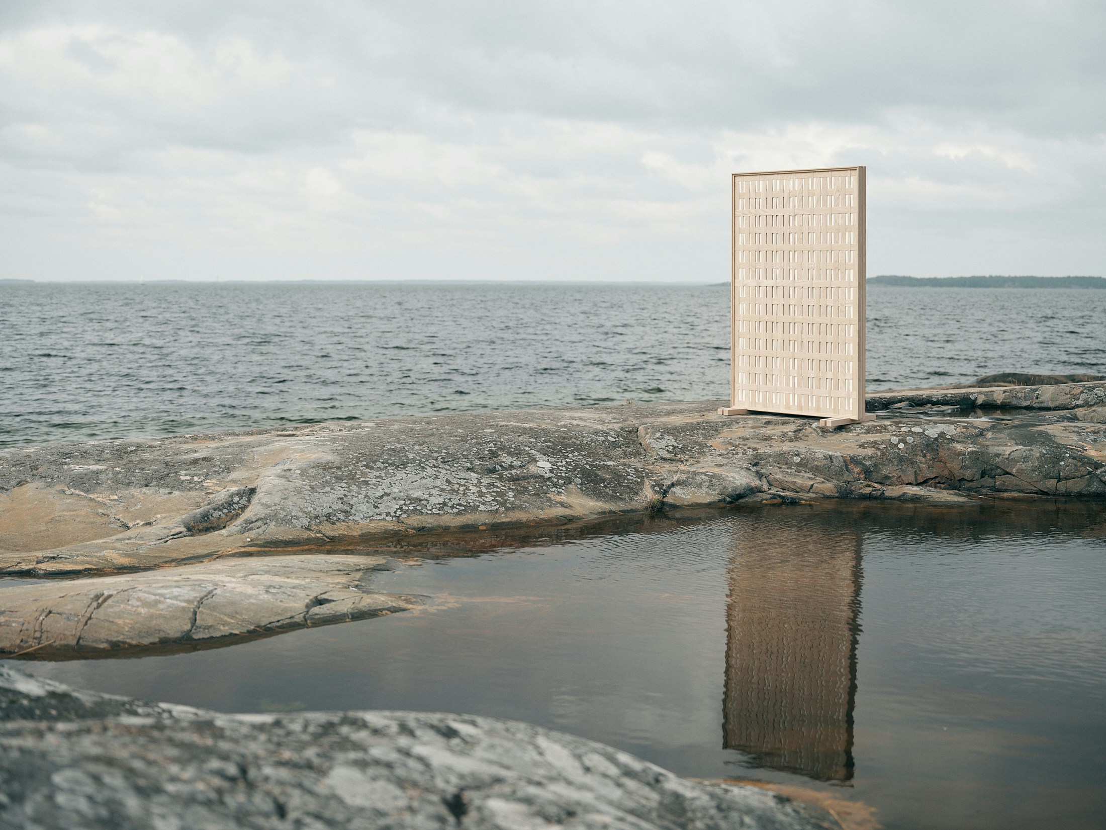 Tyyne-skærmvæggen i flettet eg står på klipperne i den finske skærgård med havet bag sig og spejler sig i en vandpyt